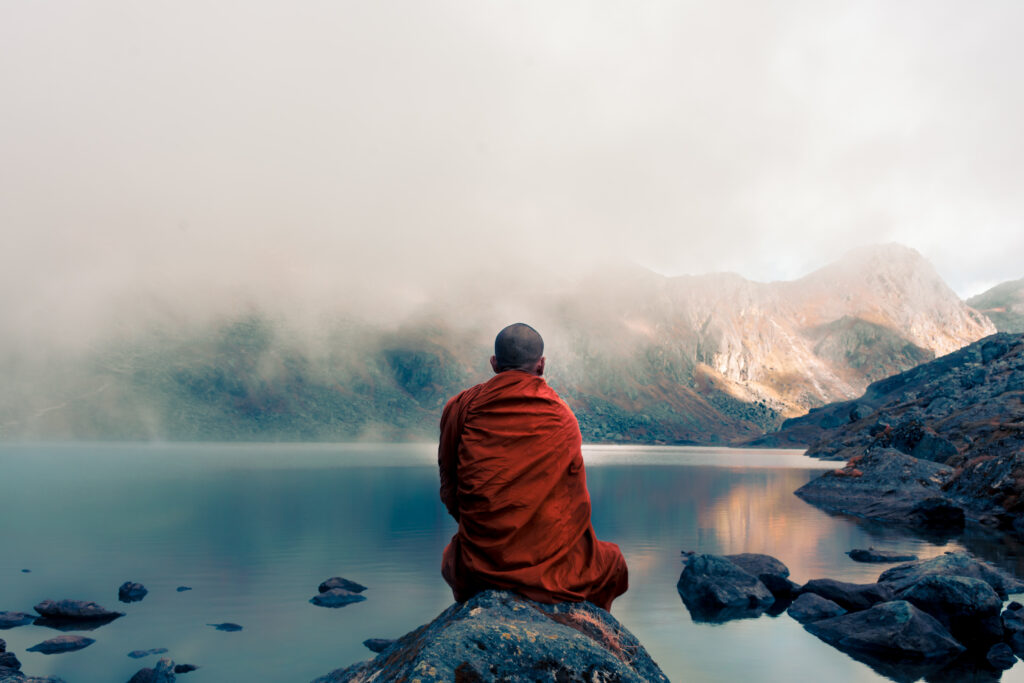 Tibetan monk from back sitting on the stone near the water in the background of foggy mountains